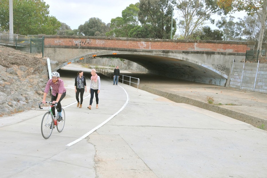 A man rides a bicycle past 2 women who are walking and engaging in coversation along a concrete path that leads under a large overheard bridge.