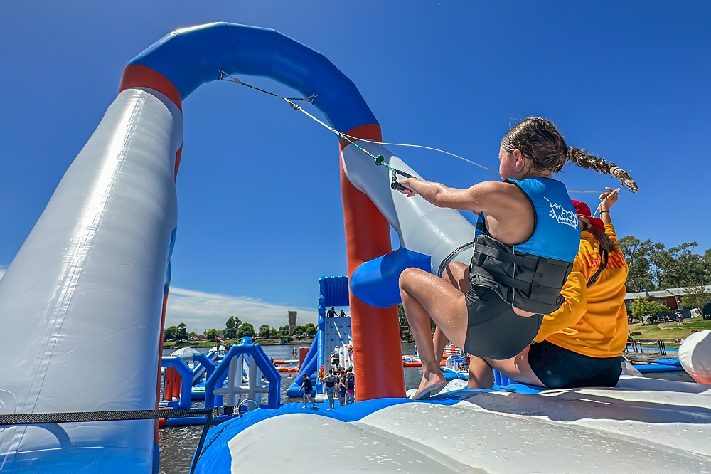 Lakes Entrance Aqua Park floating obstacles, with the back of a girl visible as she prepares to swing on a rope, with a lifeguard in a yellow jumper sitting next to her.
