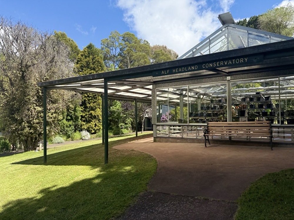 Wombat Hill’s redeveloped conservatory, with glass walls, UV protective roof and public bench all visible in the background. Various types of trees are visible to the left of the image, marking the edge of a main garden space.