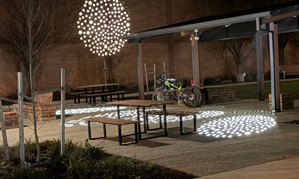 The courtyard and covered community stage outside of the Ballan Library and Community Hub, with the white light dots projected on the floor and wall. Public furniture and a green bike are visible in the image.