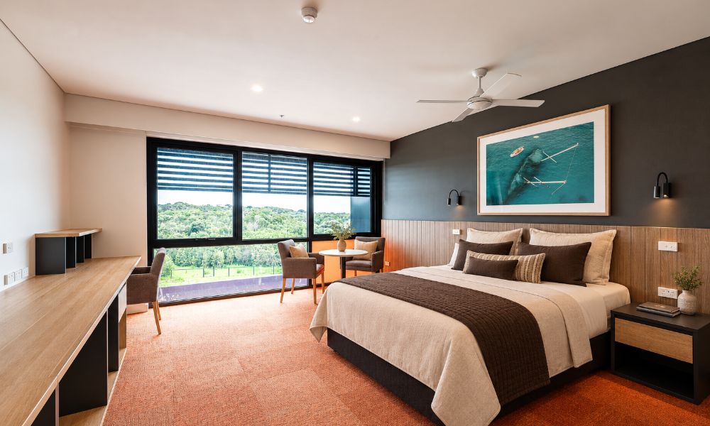  A worker apartment, featuring red carpet, a Queen Size bed, reading lamps and furniture items including a small table and long desk on the wall opposite the bed frame. A ceiling fan and framed photo of a shipwreck can be seen above the bed.