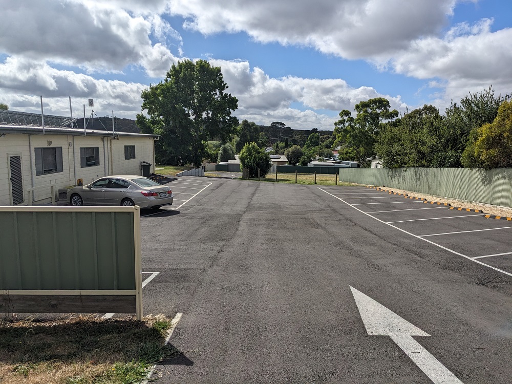 A newly built carpark with a car and a green fence on the left. There are trees in the background.  