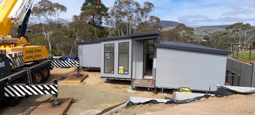 The modular house being assembled on site, with a small yellow crane visible in the front left of the picture and Omeo's rolling hills visible in the background. The modular house is made of a blue steel frame and a black steel roof.