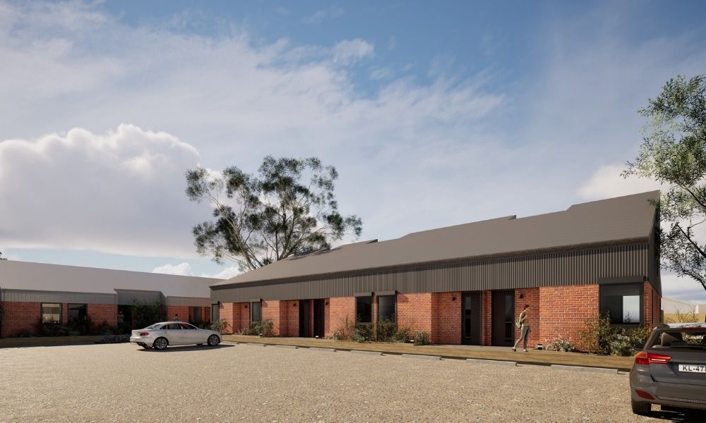 Render of the accommodation parking area, which sits in front of a brick accommodation building with a sloped black roof. A second building, with a sloped silver roof, can be seen in the back left side of the image. 