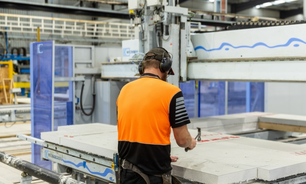 Worker in high-vis and protective gear, seen from behind, using a hammer on a section of grey woodkit that’s lying on a table-top in front of industrial machinery.