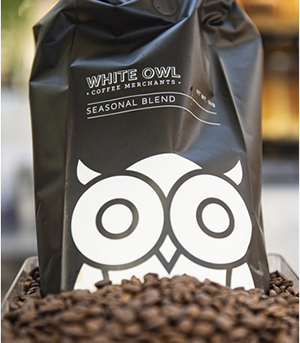 Packet of white owl coffee blend surrounded by roasted coffee beans