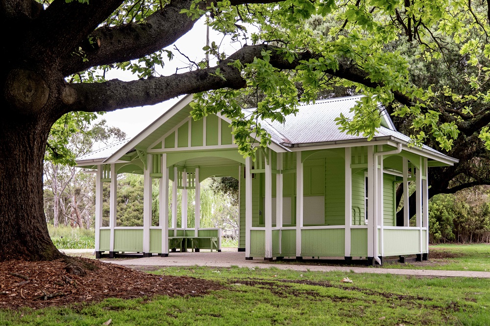 The green and white painted Montgomery Pavilion, which is framed by the branch of a large tree. In the foreground lawn is visible, with trees and garden space visible behind the pavilion in the background of the photo.