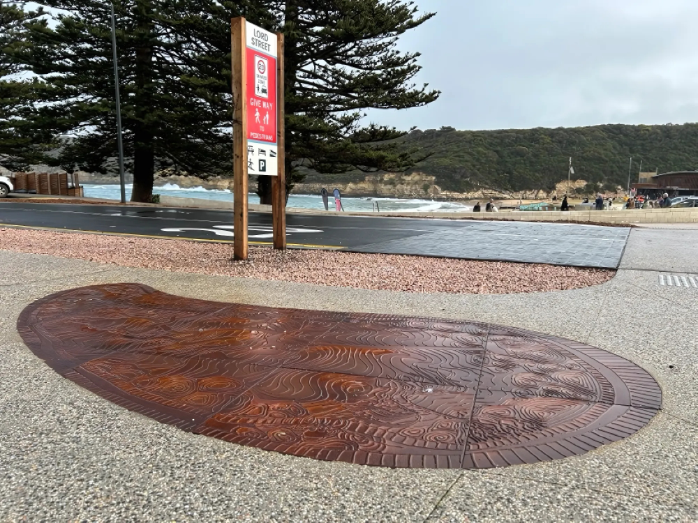Decorative First Nations art embedded into a pavement with a sign, some trees and the beach behind it. 