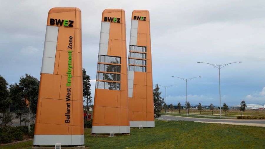 Three orange, green and silver BWEZ signs are visible from left to centre of the picture, with trees and a garden bed present behind them. To the right of the frame is a road with lamp posts and fields in the distance.