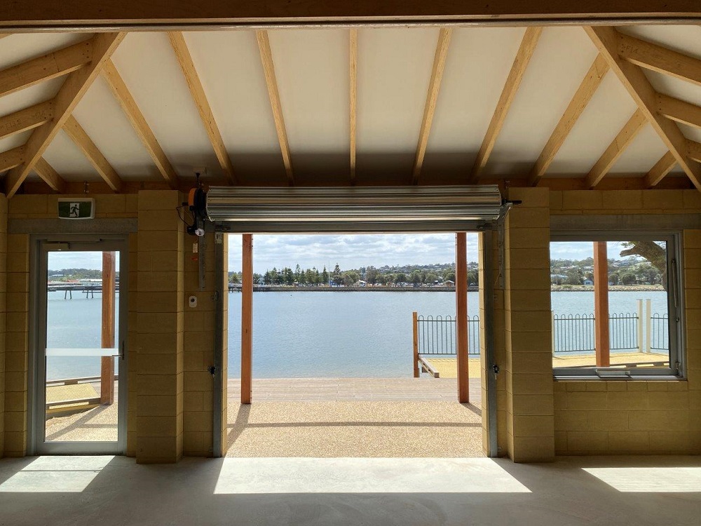 The inside of the Lakes Entrance Water Sports Pavilion, looking out on the water with the patio, lake and far shore visible out of the windows and door. 