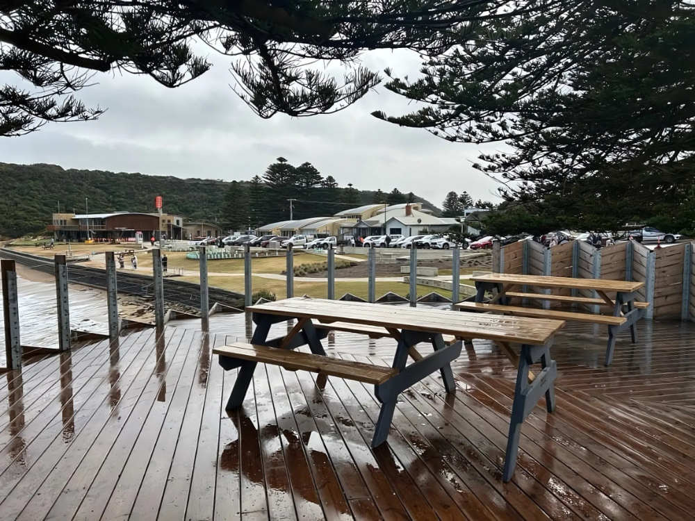 Two picnic tables on a deck, which is surrounded by a wire fence. The beach and some buildings are behind it. 
