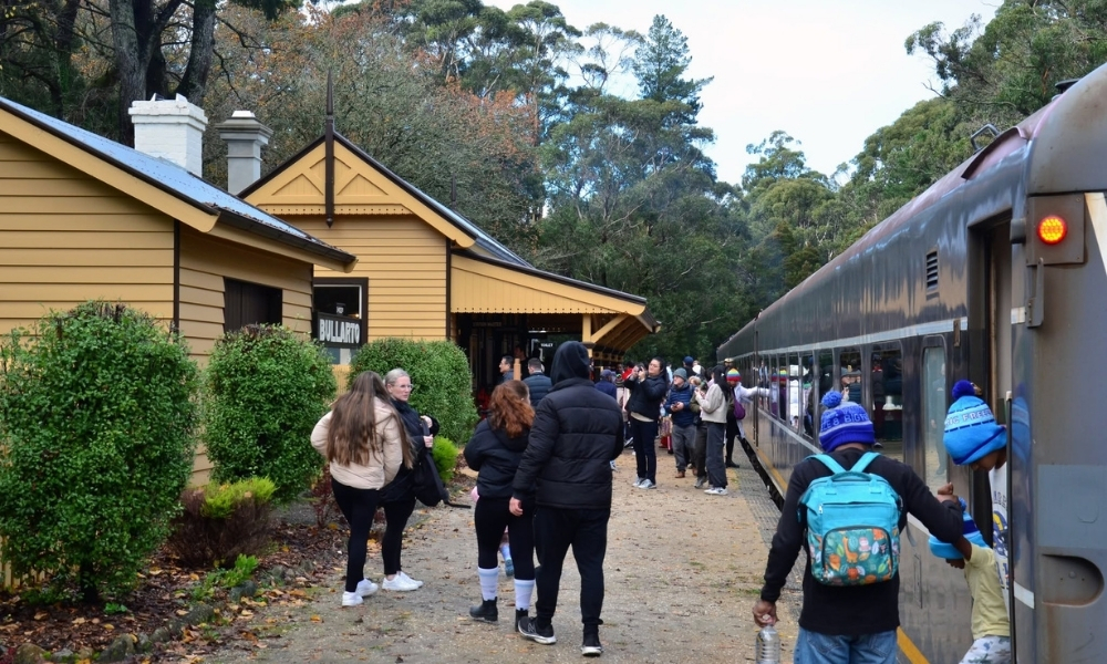 Passangers disembarking at Bullarto Station, with the historic station house visible on the left-side of the frame and the train visible to the right. The historic grounds are surrounded by tall trees and 1930’s-style shrubs and garden beds.