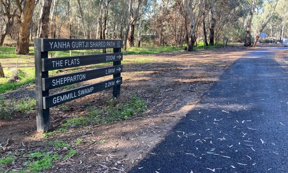 The Yanha Gurtji Shared Path, with green wooden wayfinding signage visible on the left side of the frame, with the asphalt pathway stretching into the distance on the right side of the frame.