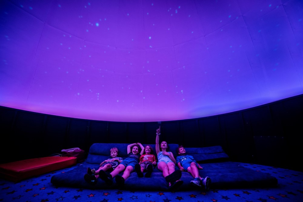 Children lay on lage cushions looking up at the domed ceiling, illuminated with hues of purple and blue with stars