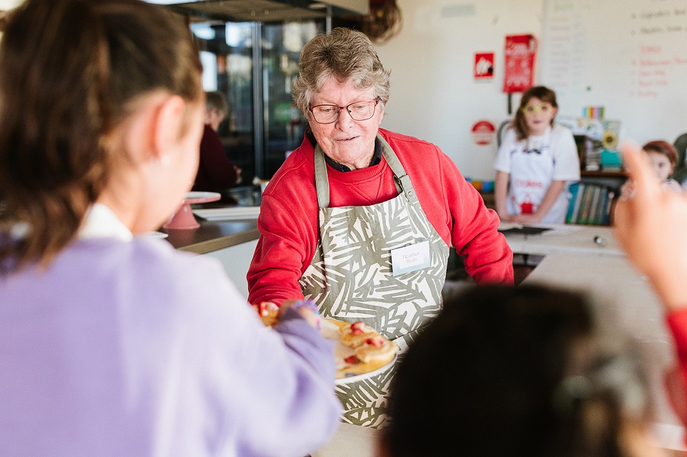 A woman in an apron and red jumper handing food to people in the foreground of the picture, using a big serving plate.