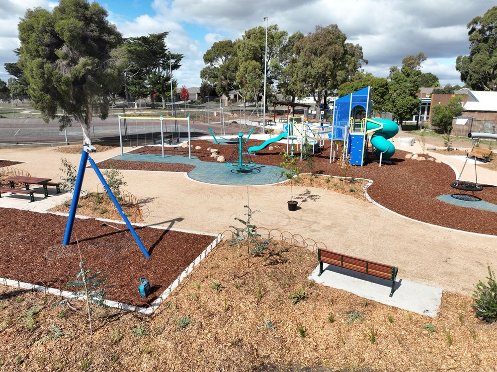 A birds-eye view of the Cardigan Village playground, with colourful equipment like slides and swings surrounded by brown tanbark and small trees. A tennis court is visible in the background.