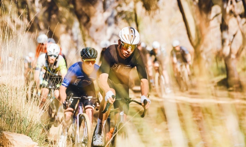 A line of gravel riders following a trail through bush and grassland, with the riders facing towards camera. Each rider is wearing different coloured safety gear, including helmets and sunglasses. 