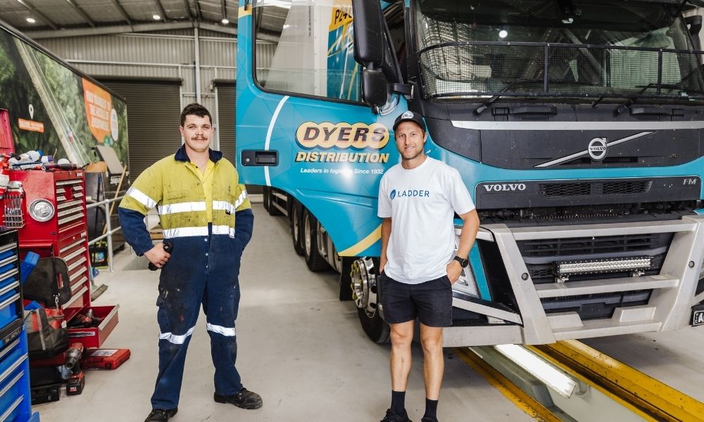 Ladder Step Up participant Jordan (R) went on to commence an apprenticeship with Dyers Distribution and became a fully qualified diesel mechanic. Ladder Step Up Program member standing in front of a Dyers Distribution truck, to the right of a worker in yellow and blue high-visibility clothing. The two men are standing in a factory with white floors and tools visible to the left of the frame.