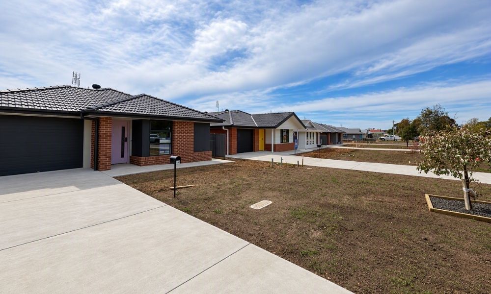 A row of brown and white worker houses, with black roofs. Each house has a concrete driveway and walkway to the front door, and a front lawn area with a tree planted near the edge of the property.