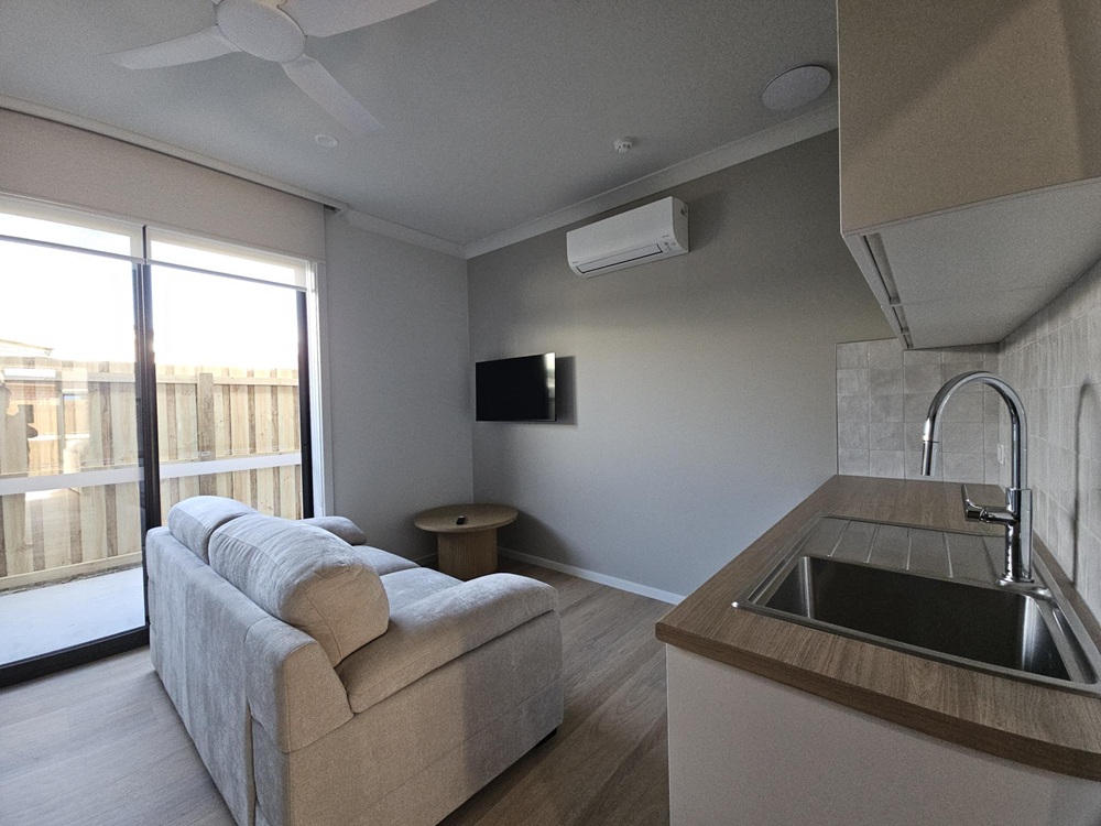 The interior of one of the seven units, showing the living room area complete with a kitchenet and wet bar, with white split system air conditioner unit and a black TV mounted to the wall.  