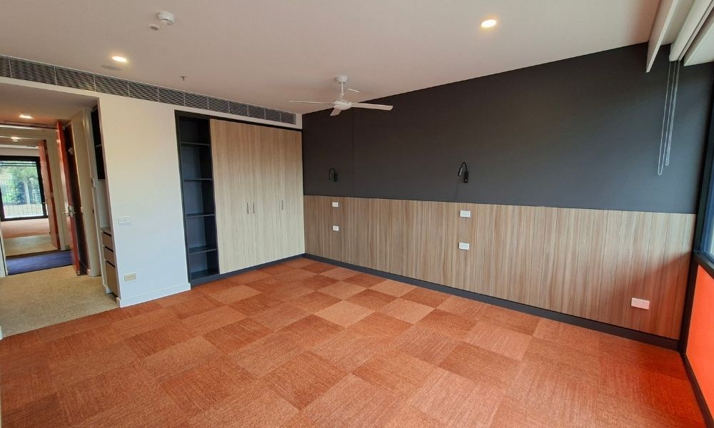 The interior of one of the accommodation units, showing wooden wall panelling, a white ceiling with a fan, chequered carpet and an entrance hallway out into the shared space.