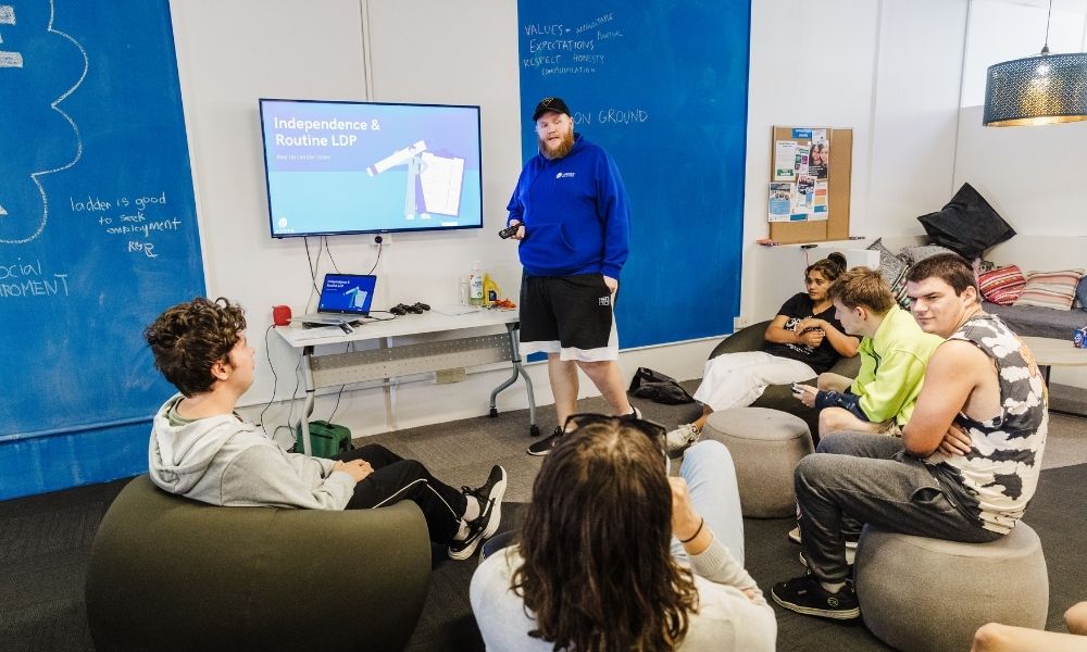 Ladder Step Up is deliberately designed to engage young people in an environment that feels very different from a classroom – this session focused on independence and building practical skills. Ladder Step Up Program leader standing in front of a tv and blue walls, speaking to several youth who are seated on beanbag chairs and listening to his presentation.