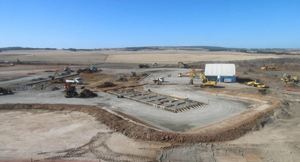 Early works on the Visitor Experience Centre are visible at the centre of the frame, with concrete slabs, mounds of soil and various construction vehicles visible from an elevated viewpoint. The background of the image shows rolling hills and fields, with some greenery visible just under the horizon