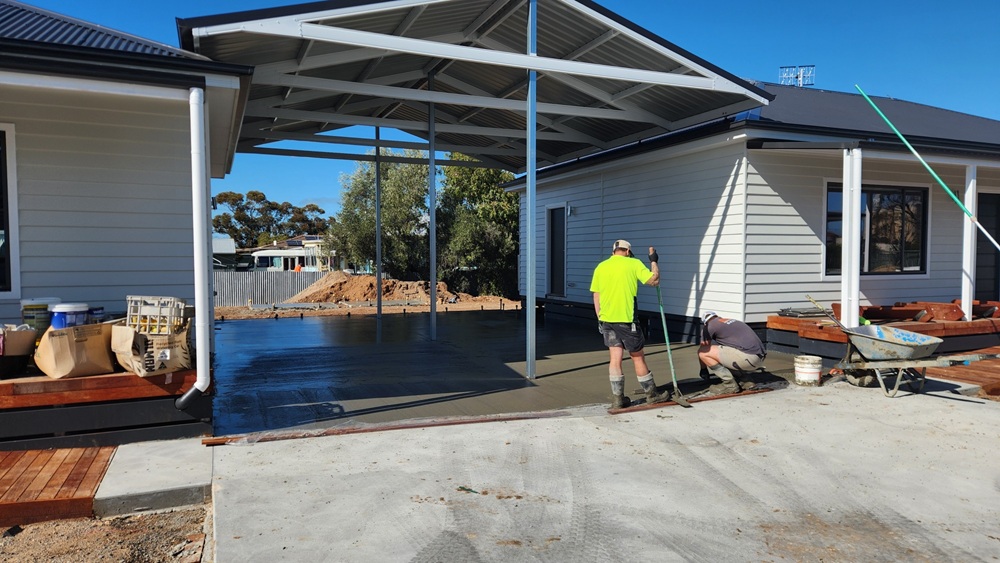 Two tradies laying the concrete for the shared carport between the two duplexes. One tradie is standing to the left, leaning on a broom, while the second tradie is squatting to the right side, working on the wet concrete. A carport roof is centre frame, with the two duplexes visible on either side.