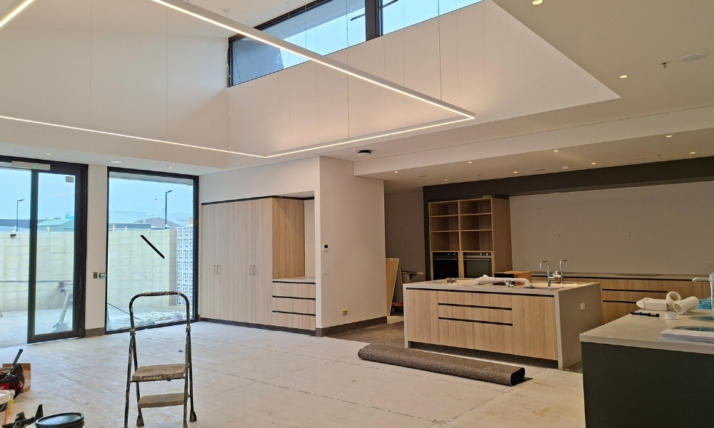 The accommodation's shared public space, with an open plan kitchen area, storage and living space. The walls and floor are both white, and construction tools and a step ladder are visible in the image.