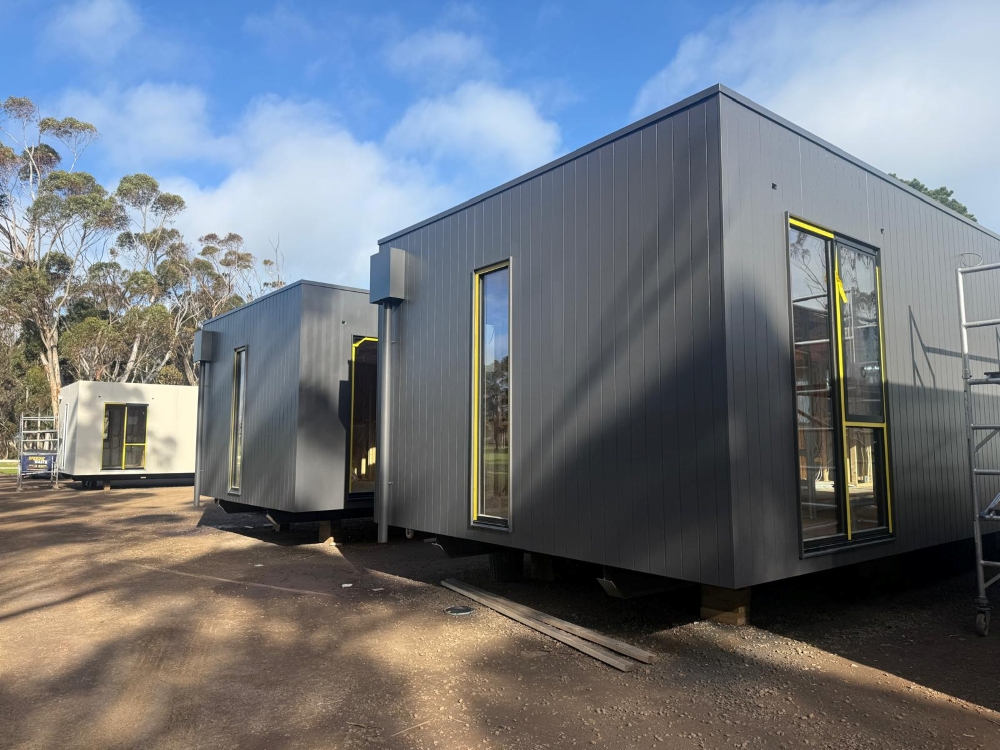 Modular housing units under construction, with grey cladding and large windows, positioned on a gravel site near bushland.