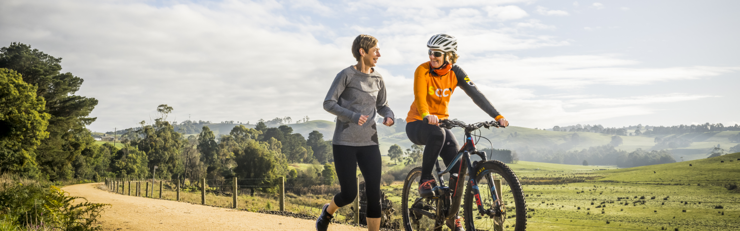 A woman iides a bike while chatting with a friend who jogs beside her. They are travelling along a gravel road in a lush and colourful countryside location