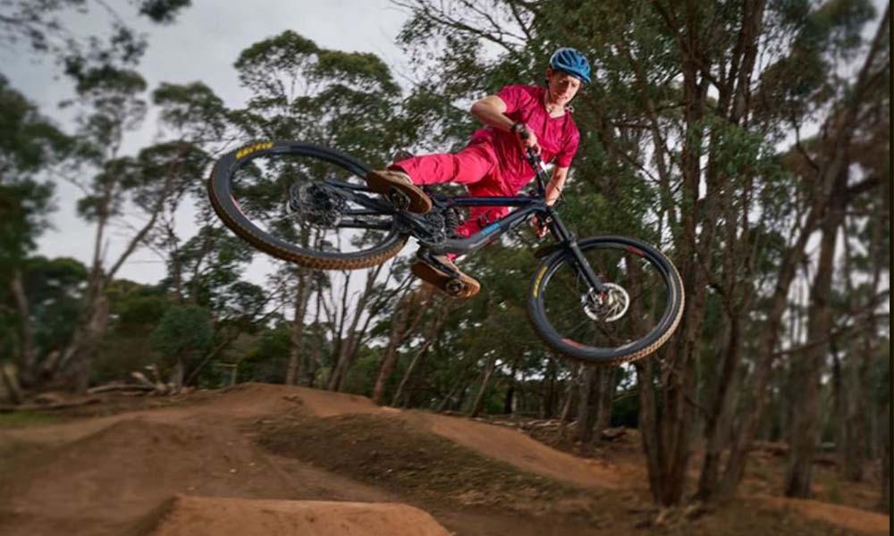 Rider in the centre of image is in the air between two jumps, wearing a red riding suit and blue helmet, with their bike angled to the right. The bottom of the image shows dirt BMX ramps, while the background of the image shows gum trees surrounding the trail. 