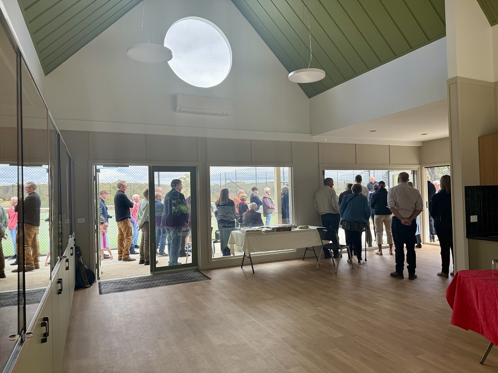 People gathered at the newly opened Navigators Hub. In the foreground you can see hardwood floors, wall-mounted glass cabinets and display cases and a red and white table to cater for a function. People are seen listening to a speaker at the edge of the building and outside of the building and are visible through the open doors and clear windows.