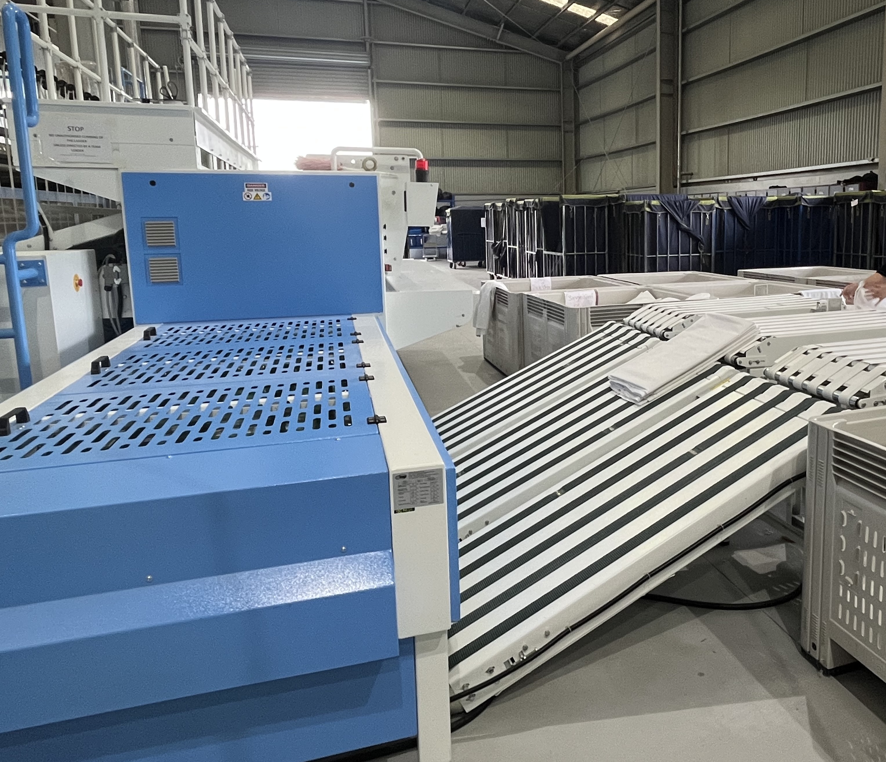Surf Coast Laundry has received equipment upgrades to boost output and jobs. Large blue-and-white industrial laundry machine with a roller conveyor system inside a warehouse.