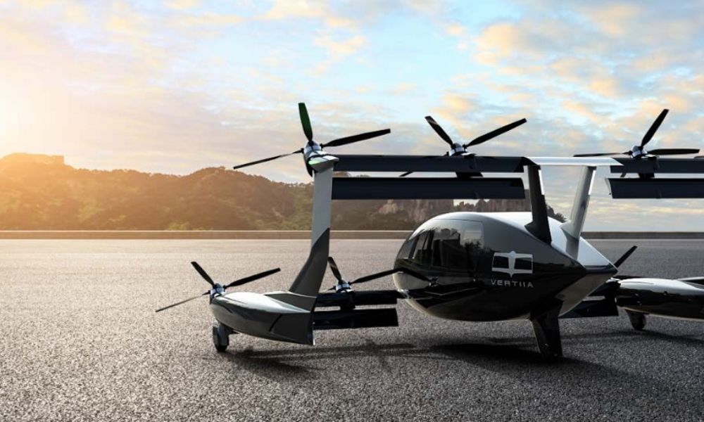 A black propeller drone sitting on concrete, facing forested hills with the sun rising over them.
