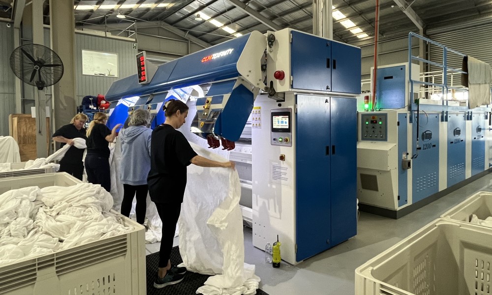 Workers at laundry holding sheets towards a large machine.