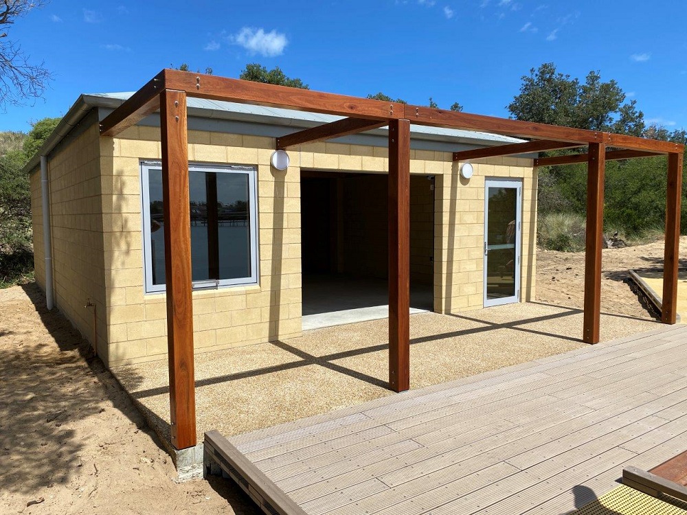 The Lakes Entrance Water Sports Pavilion, a yellow limestone single-story building, with wooden patio framing at the front of the building and decking in front of that, leading to the beach.