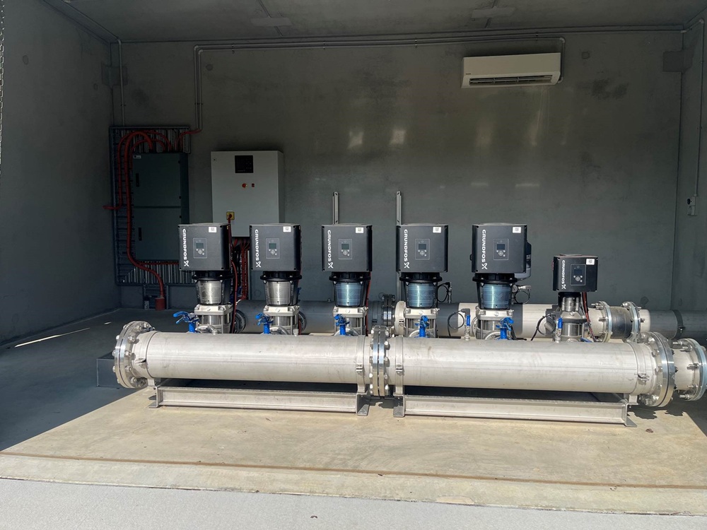 Six black electronic water pumps, lined up from left to right behind a grey steel pipe.