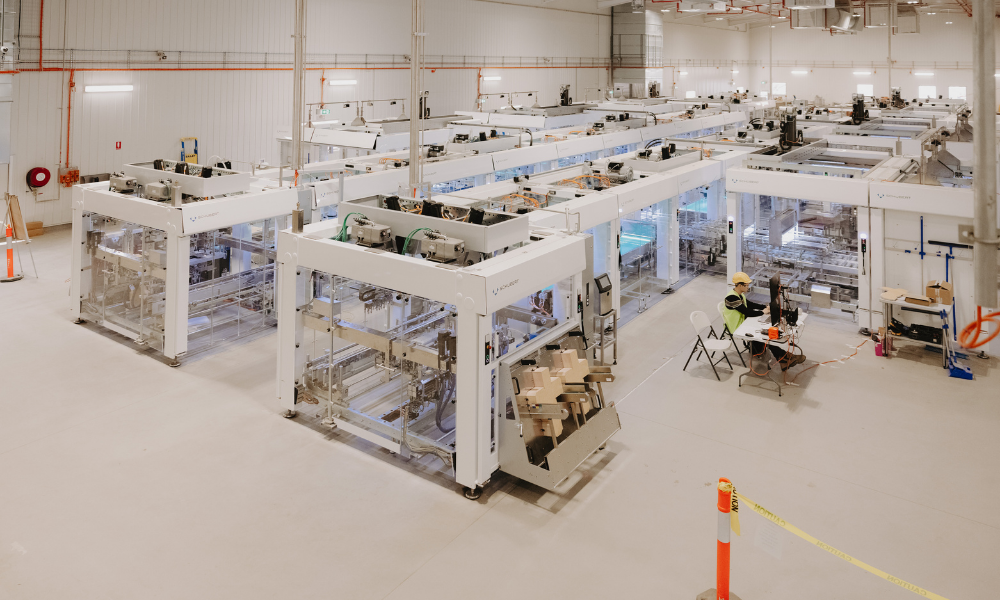 White factory floor with pod-style manufacturing infrastructure made from metal and glass. Assembly machinery can be seen through the glass.