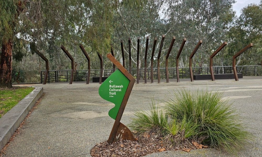 A brown wood and green metal sign pointing to the Bullawah Cultural Trail, with the Torryong (Ovens) riverfront trail entrance visible in the back of the image.