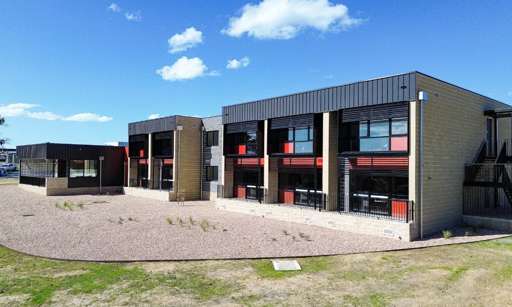 The exterior of the Bayside Health worker accommodation building. The building is two stories high, with a grey brick exterior, red and black finish on the outside of the windowed apartments, which all have a black roof. A native garden and lawn area is visible in front of the building.