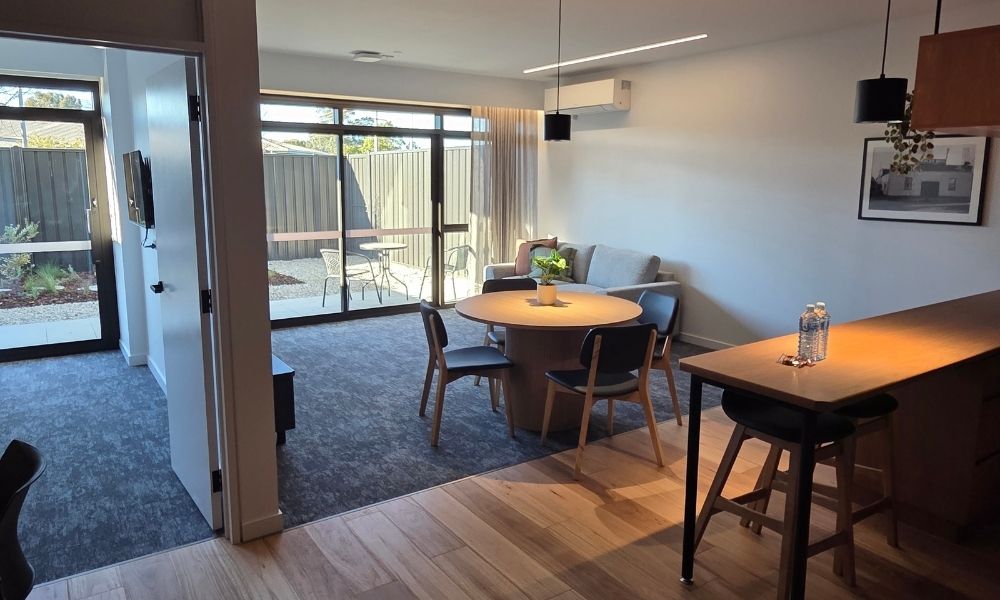 Inside one of the GV Health units, which contain a kitchenette with a hardwood floor, and carpeted living and bedroom spaces that look out into a fenced balcony or courtyard.