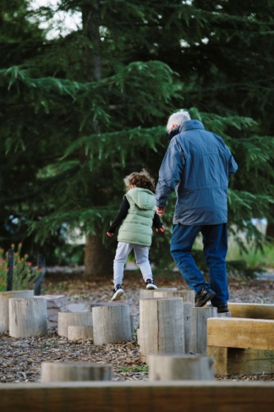 A grandfather and granddaughter walking and balancing on timber logs at the new Romsey Ecotherapy Park with Pine trees in the background