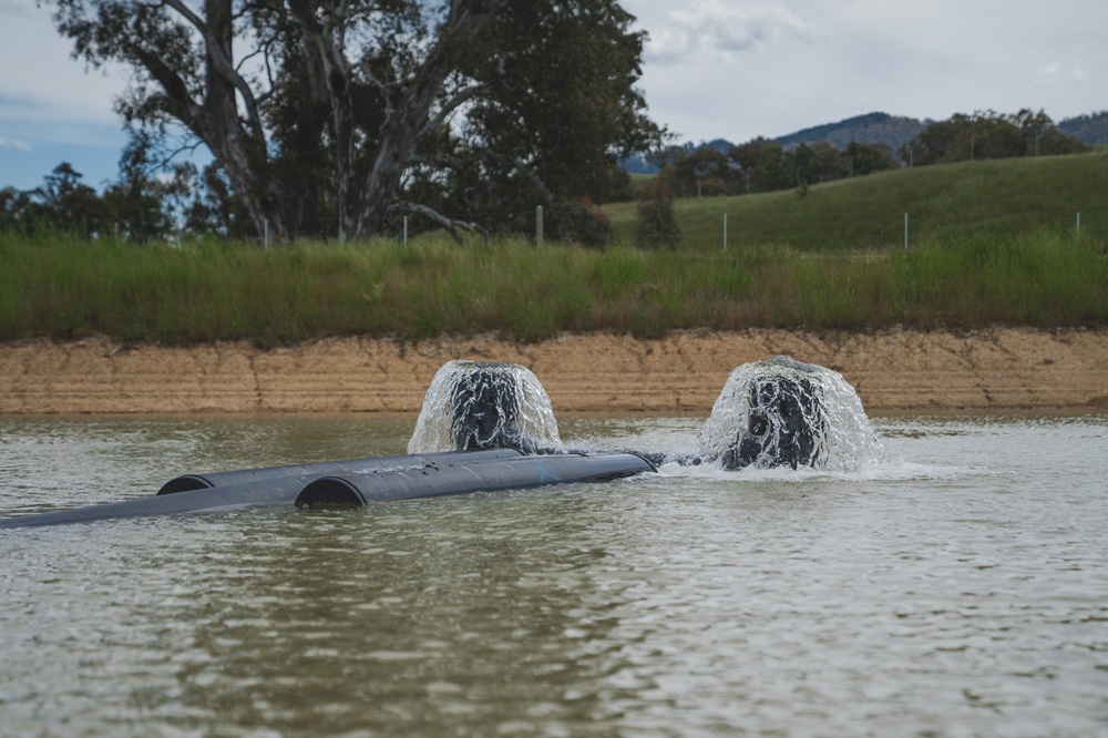 Black water pipeline pushing water out through two vertical nozzles, creating a fountain effect. The surrounding water is brown, and green grass, gum trees and hills can be seen to the top of the frame.