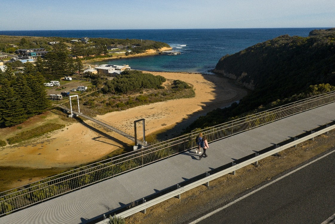 A scenic coastal view featuring a pedestrian bridge spanning a sandy inlet, connecting two elevated green areas.