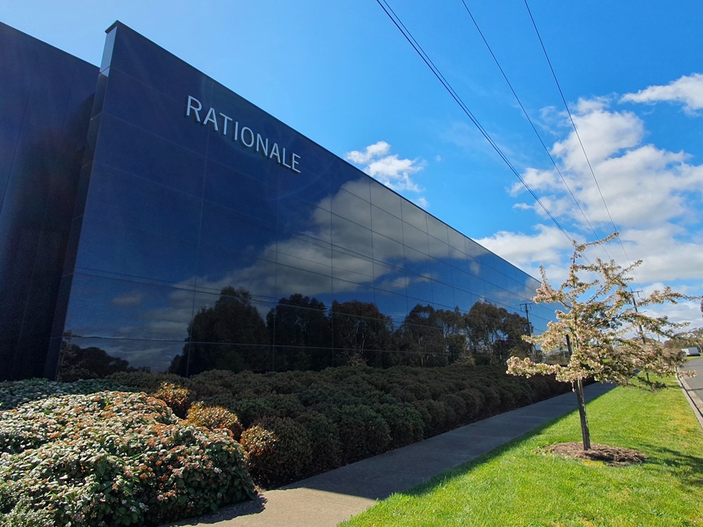 A building with black windows reflecting trees. The word, “RATIONALE” is on it. There are also bushes, a sidewalk and a nature strip with floral trees in front of it.