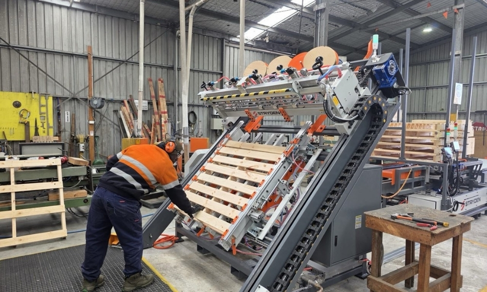 Worker operating an automated pallet manufacturing machine inside a timber production facility, with completed pallets visible in the background.