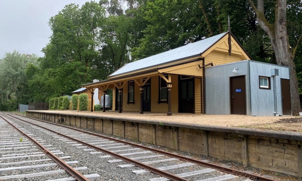 Two railway lines in front of Bullarto Station platform, a small historic building with a tin toilet block at its side. Tall trees cover the background of the station. 
