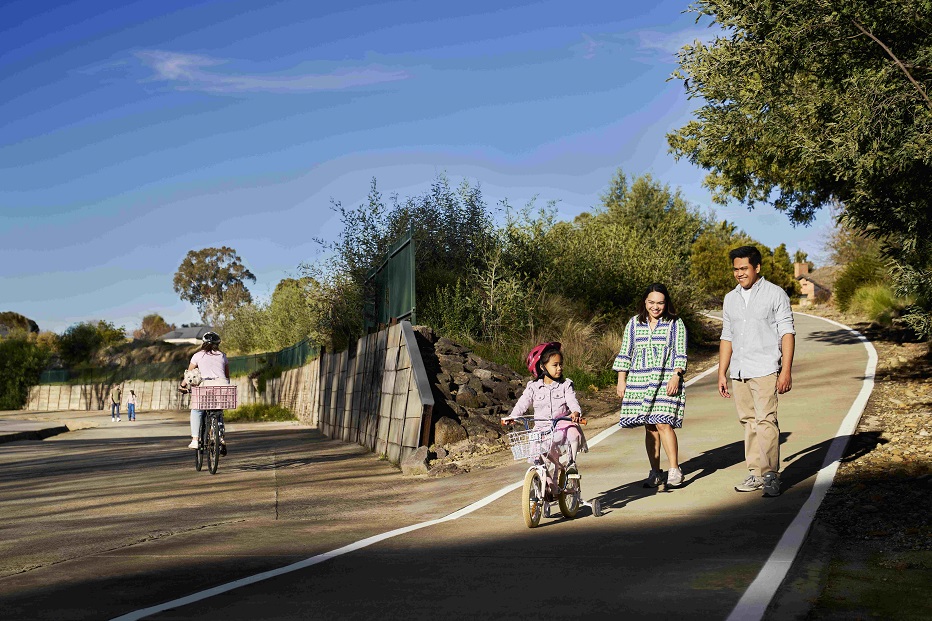 A mother and father walk with their young daughter riding a bike along a concrete path with trees surrounding the area