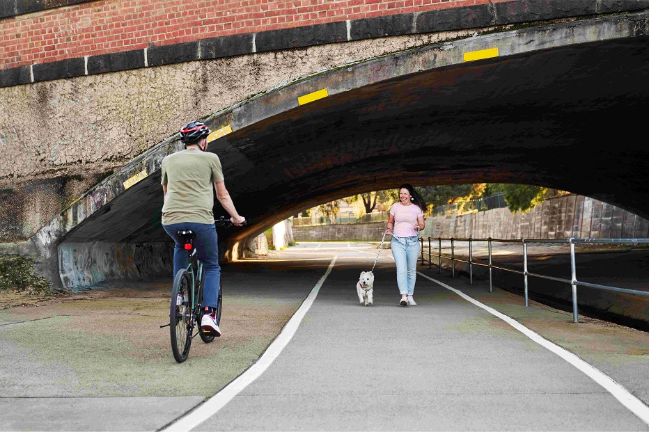 A man rides a bicycle past a woman walking her dog along a concrete path that leads under a large overheard bridge.